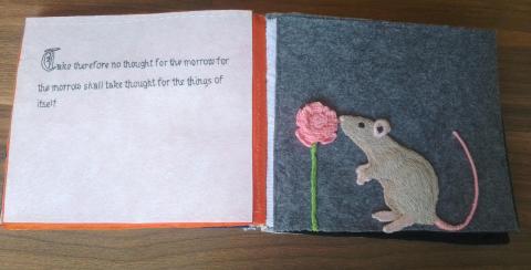 The text is to the left, embroidered in black thread on a white background, and the rat is to the right, on a grey background